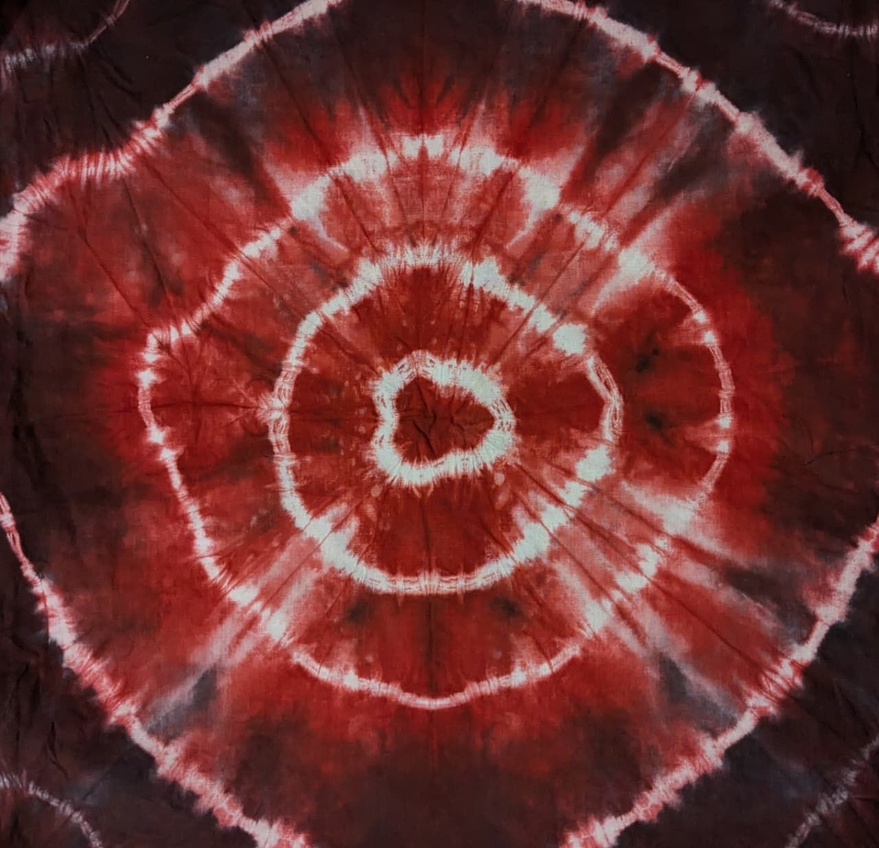 Large circular tie-dye medallion in deep maroon and red on dark cloth, with concentric rings of resist patterning